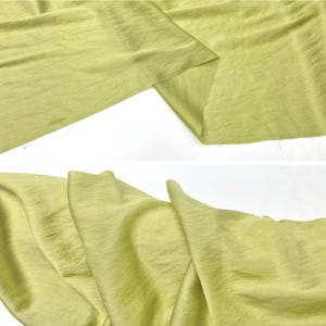 May include: A length of light lime green fabric with a subtle, fine-grained texture. The material is shown in two sections, displaying soft, flowing folds and a matte finish that catches the light across its surface, highlighting the weave.