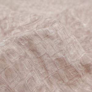 May include: Close-up of a folded piece of fabric in a light beige color. The fabric has a textured surface with a repeating pattern of small, square-like shapes. The fabric is soft and appears to be made of a delicate material.