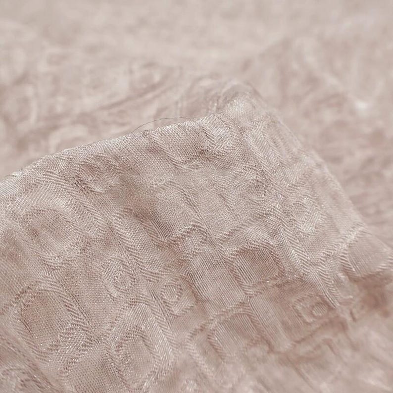May include: Close-up of a folded piece of fabric in a light beige color. The fabric has a textured surface with a repeating pattern of small, square-like shapes. The fabric is soft and appears to be made of a delicate material.