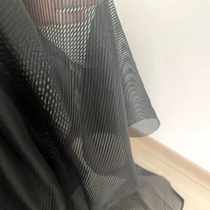 May include: Close-up of a sheer black fabric with a striped pattern. The fabric is draped, revealing a glimpse of the background. The material appears lightweight and has a subtle sheen, suggesting it could be used for clothing or decorative purposes.