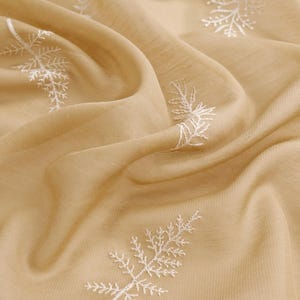 May include: A close-up of a beige fabric with white embroidered floral patterns. The fabric is draped, creating soft folds and shadows. The embroidery is detailed, with delicate leaf-like designs. The overall texture appears soft and lightweight.