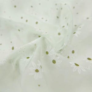 May include: A light sage green fabric featuring a repeating pattern of white daisy flowers with yellow centers. The material has a soft, semi-sheer texture with gentle folds and drapes that create a sense of depth across the surface of the textile.