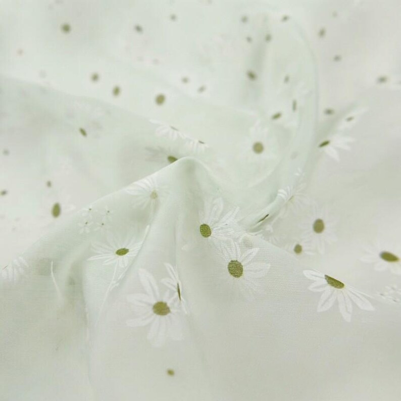 May include: A light sage green fabric featuring a repeating pattern of white daisy flowers with yellow centers. The material has a soft, semi-sheer texture with gentle folds and drapes that create a sense of depth across the surface of the textile.