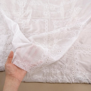 Op de afbeelding: Witte eyelet stof met gedetailleerde bloemen en geometrische borduursels. Het dunne materiaal is gevouwen en toont het gedetailleerde openwerk ontwerp. De stof is lichtgewicht, waarschijnlijk katoen of een katoenmix. Het patroon is zichtbaar.