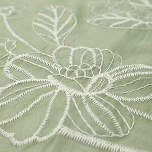 May include: A sage green fabric featuring white thread embroidery in a floral pattern. The design consists of large, outlined flower blossoms and leaves stitched directly onto the semi-sheer, lightweight textile surface with visible thread texture.