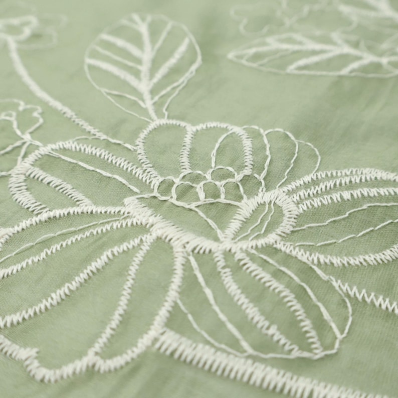 May include: A sage green fabric featuring white thread embroidery in a floral pattern. The design consists of large, outlined flower blossoms and leaves stitched directly onto the semi-sheer, lightweight textile surface with visible thread texture.