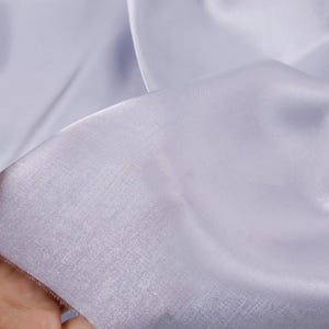 May include: Close-up of a light lavender fabric with a soft, flowing texture. The fabric appears to be sheer, with subtle light reflections. The material is likely used for apparel or decorative purposes.
