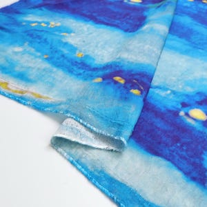 May include: A folded textile with a watercolor-style design. The fabric features shades of blue, with lighter areas and darker, more saturated sections. Small yellow accents are scattered across the surface. The edge of the fabric is visible, showing a white, textured underside.