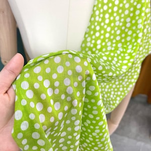 May include: A lightweight fabric scarf featuring a vibrant lime green background with a pattern of white polka dots of varying sizes. The material has a visible weave texture and is draped to show the repeating circular design across the surface.
