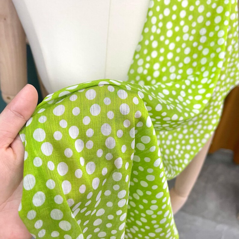 May include: A lightweight fabric scarf featuring a vibrant lime green background with a pattern of white polka dots of varying sizes. The material has a visible weave texture and is draped to show the repeating circular design across the surface.