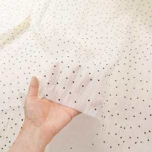 May include: Off-white fabric with a delicate, sheer texture, scattered with small, evenly spaced black dots. The fabric is held up, revealing its lightweight quality and the pattern's subtle elegance. Ideal for apparel or home decor.