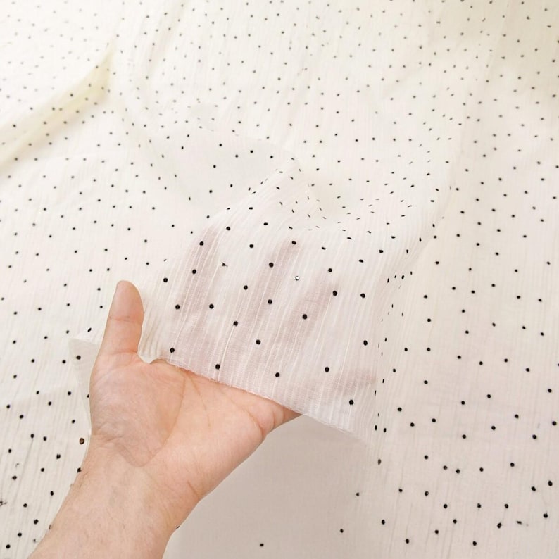 May include: Off-white fabric with a delicate, sheer texture, scattered with small, evenly spaced black dots. The fabric is held up, revealing its lightweight quality and the pattern's subtle elegance. Ideal for apparel or home decor.