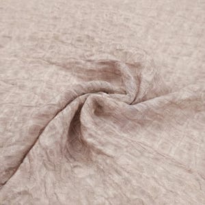 May include: A close-up shot of a light mauve fabric with a textured, crinkled surface. The fabric is gathered and folded, creating soft shadows and highlights. The material appears to be lightweight and flowing, suitable for apparel or home decor.