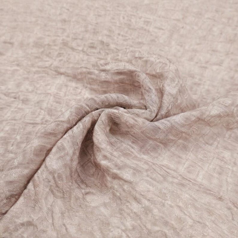 May include: A close-up shot of a light mauve fabric with a textured, crinkled surface. The fabric is gathered and folded, creating soft shadows and highlights. The material appears to be lightweight and flowing, suitable for apparel or home decor.
