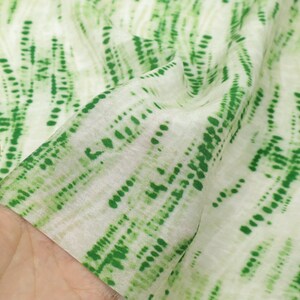 Op de afbeelding: Een lichtgewicht stof met een witte basis en een herhalend verticaal patroon van groene, gestreepte en gestippelde lijnen. Het materiaal heeft een zachte, semi-transparante textuur met zichtbare plooien die diepte en beweging in de stof cre&euml;ren.