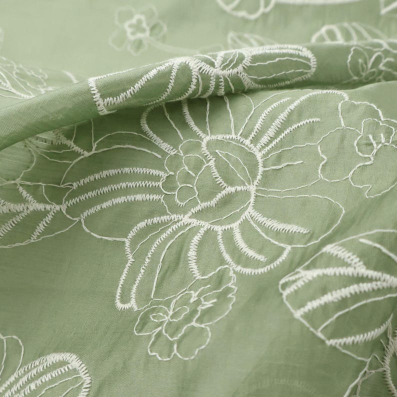 May include: A sage green fabric featuring a delicate white embroidered floral pattern. The embroidery consists of thin, continuous lines forming large, stylized blossoms and leaves across the surface of the lightweight, slightly translucent textile.