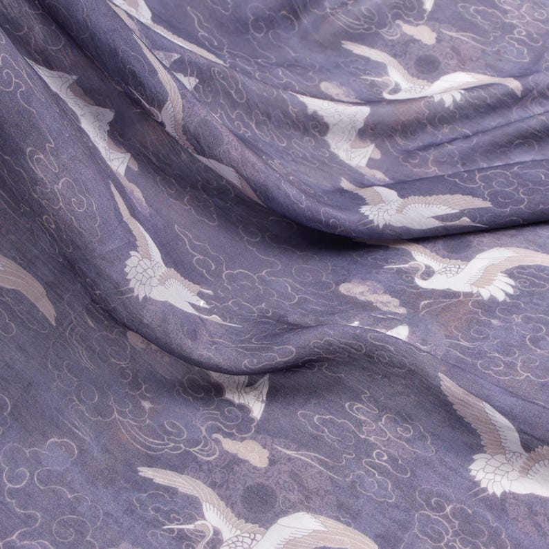May include: Close-up of a fabric with a repeating pattern of white cranes and cloud motifs on a muted purple background. The fabric is draped, showing folds and texture. The design evokes a sense of elegance and tranquility.