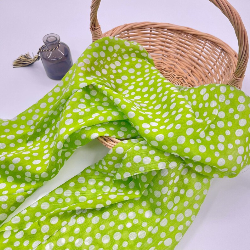 May include: A bright lime green scarf featuring a white polka dot pattern draped over a light brown woven wicker basket. A small, dark gray glass bottle with a decorative tassel is positioned to the left of the basket on a white surface.