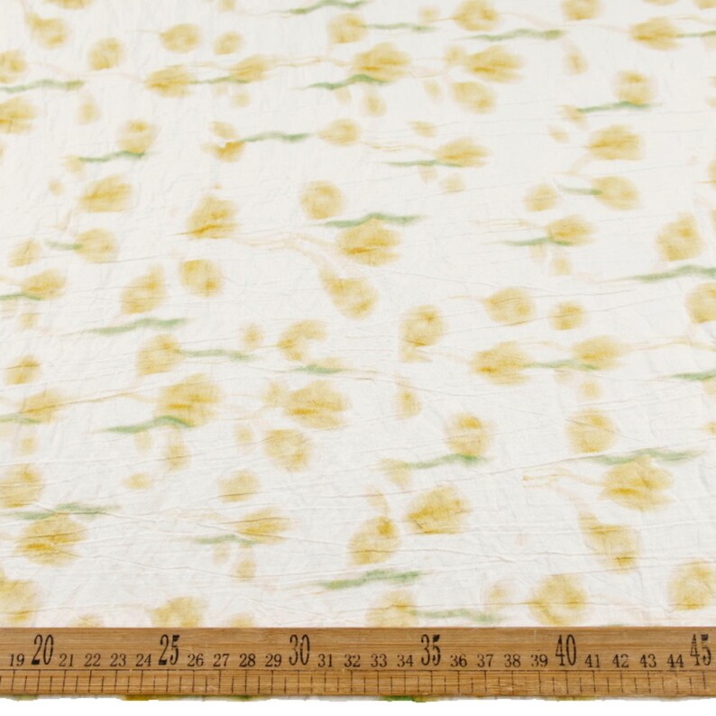May include: A white fabric with a crinkled texture featuring a repeating pattern of soft yellow floral shapes and light green stems. A wooden ruler is positioned at the bottom, showing measurements from 19 to 45 inches across the fabric width.
