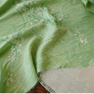 Peut inclure: Un tissu vert clair avec un motif floral de fleurs blanches et de tiges vertes. Le tissu a un aspect l&eacute;g&egrave;rement textur&eacute; et est drap&eacute;, r&eacute;v&eacute;lant un tissu blanc contrastant avec un motif floral en dessous.