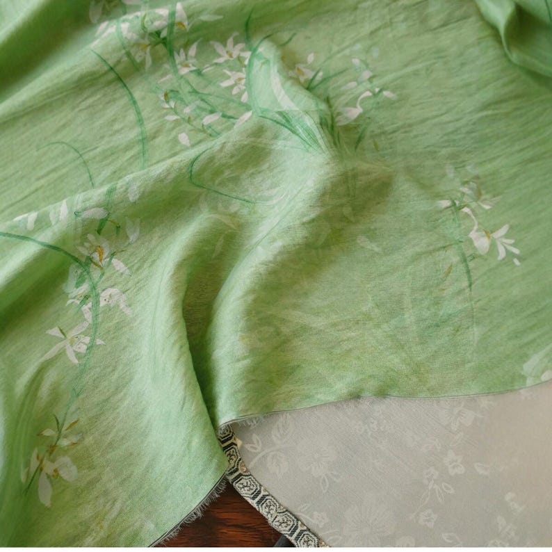 Peut inclure: Un tissu vert clair avec un motif floral de fleurs blanches et de tiges vertes. Le tissu a un aspect l&eacute;g&egrave;rement textur&eacute; et est drap&eacute;, r&eacute;v&eacute;lant un tissu blanc contrastant avec un motif floral en dessous.