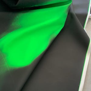 Peut inclure: Un rouleau de tissu présentant une surface extérieure noir mat qui se replie pour révéler une doublure intérieure vert néon vif. Le matériau semble lisse et flexible, la couleur vert vif créant un contraste marqué avec le noir foncé.