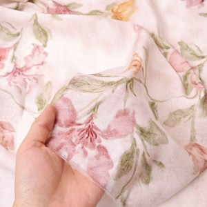 May include: A delicate, floral fabric with a watercolor-style design. The fabric features pink, peach, and yellow flowers with green leaves on a white background. The material is soft and flowing, ideal for scarves or dresses.