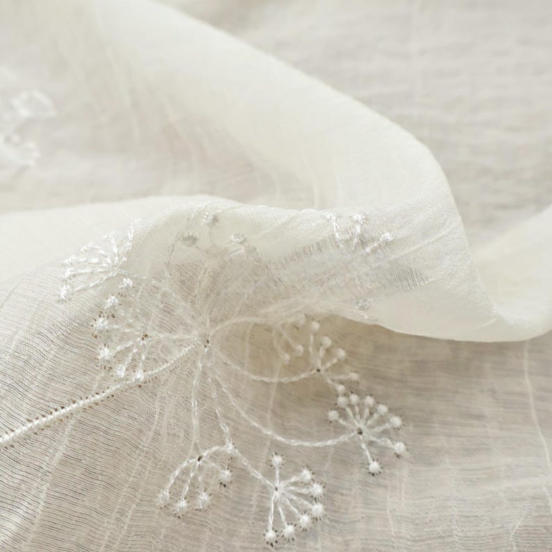 May include: Close-up of a sheer, off-white fabric with delicate floral embroidery. The embroidery features a detailed, airy design with small, beaded accents. The fabric is gently draped, showcasing its texture and the intricate details of the needlework.