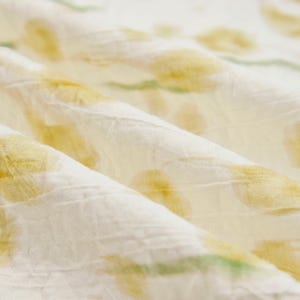 May include: A white fabric with a crinkled texture featuring a repeating pattern of soft yellow floral shapes and light green stems. The material is folded, creating deep ridges and shadows that highlight the textured surface of the textile.