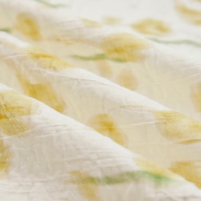 May include: A white fabric with a crinkled texture featuring a repeating pattern of soft yellow floral shapes and light green stems. The material is folded, creating deep ridges and shadows that highlight the textured surface of the textile.