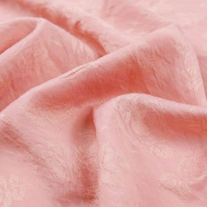 May include: A soft, light pink fabric featuring a tonal, woven floral jacquard pattern. The material is draped in gentle folds, highlighting the subtle texture and the repeating rose motif integrated directly into the weave of the textile.