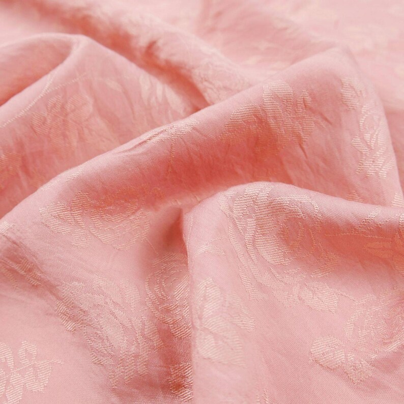 May include: A soft, light pink fabric featuring a tonal, woven floral jacquard pattern. The material is draped in gentle folds, highlighting the subtle texture and the repeating rose motif integrated directly into the weave of the textile.