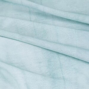 K&ouml;nnte beinhalten: Nahaufnahme eines weichen, hellblauen Stoffes mit subtilen Farbvariationen. Das Material scheint drapiert zu sein, mit sanften Falten und Schatten, die Tiefe und Textur erzeugen. Der Gesamteindruck ist weich und leicht.