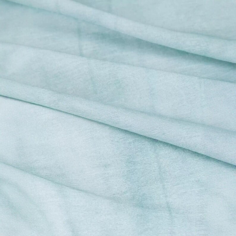 K&ouml;nnte beinhalten: Nahaufnahme eines weichen, hellblauen Stoffes mit subtilen Farbvariationen. Das Material scheint drapiert zu sein, mit sanften Falten und Schatten, die Tiefe und Textur erzeugen. Der Gesamteindruck ist weich und leicht.
