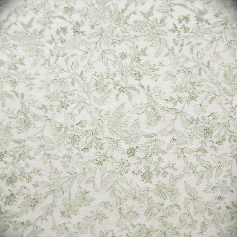 Puede incluir: Primer plano de una tela con un patr&oacute;n floral repetido. El dise&ntilde;o presenta hojas y flores de color verde claro sobre un fondo blanco. El patr&oacute;n es detallado y cubre toda la superficie.