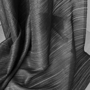 Puede incluir: Una tela de color gris carbón oscuro que presenta una textura plisada vertical fina. El material está drapeado en pliegues suaves y fluidos, creando una serie de patrones de luz y sombra en la superficie que resaltan la construcción lineal y acanalada.