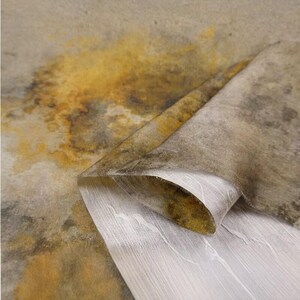 Peut inclure: Une feuille de mat&eacute;riau textur&eacute; pr&eacute;sentant un motif tachet&eacute; dans des tons terreux de brun, d'ocre et de gris. La couche sup&eacute;rieure est repli&eacute;e pour r&eacute;v&eacute;ler un envers blanc, fibreux et lisse, avec un grain lin&eacute;aire subtil et translucide.