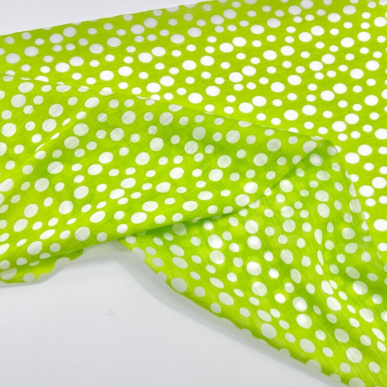 May include: A bright lime green fabric featuring a repeating pattern of white polka dots of varying sizes. The material has a subtle, fine-textured, linear weave visible across the surface, creating a light-catching effect on the fabric.