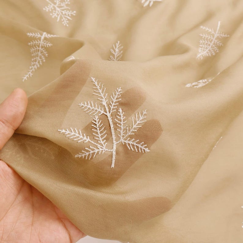 May include: Tan sheer fabric with white embroidered leaf patterns. The fabric is held up, showing its delicate, translucent quality. The embroidery features detailed, fern-like designs, creating a natural, elegant aesthetic.