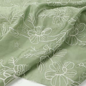 May include: A sage green fabric featuring a white embroidered floral pattern. The design consists of large, detailed blossoms and leaves stitched in white thread across the surface of the light green textile, creating a textured appearance.
