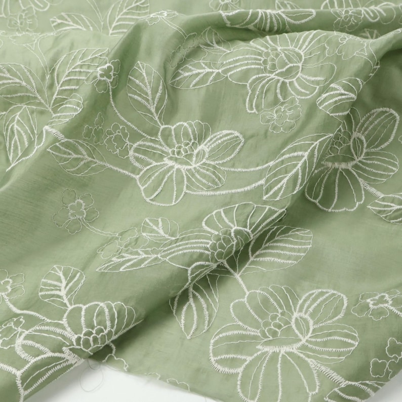 May include: A sage green fabric featuring a white embroidered floral pattern. The design consists of large, detailed blossoms and leaves stitched in white thread across the surface of the light green textile, creating a textured appearance.