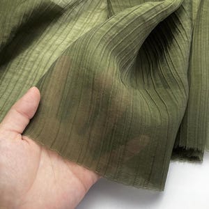 May include: Close-up of olive green fabric with vertical ribbed texture. The sheer material is held up, revealing its delicate nature. The fabric has a subtle sheen and a soft, flowing appearance, suitable for apparel or home decor.
