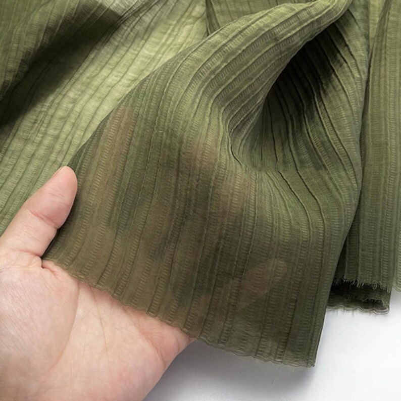 May include: Close-up of olive green fabric with vertical ribbed texture. The sheer material is held up, revealing its delicate nature. The fabric has a subtle sheen and a soft, flowing appearance, suitable for apparel or home decor.
