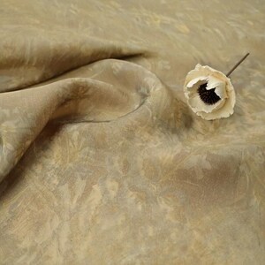 May include: Close-up of a beige fabric with a subtle floral pattern. The fabric is draped, creating soft folds and shadows. A single, off-white flower with a dark center rests on the fabric, adding a touch of contrast.