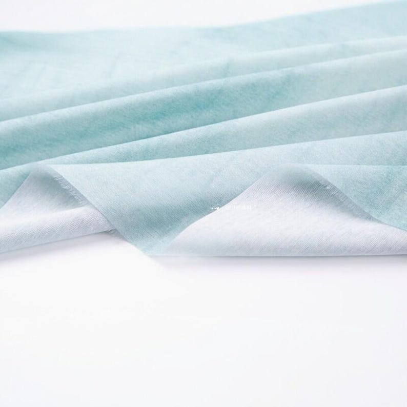 K&ouml;nnte beinhalten: Nahaufnahme eines hellblauen Stoffes mit Aquarelleffekt. Der Stoff ist gefaltet und drapiert und zeigt eine weiche Textur und einen Farbverlauf von Wei&szlig; zu Hellblau. Der Stoff wirkt leicht und luftig.