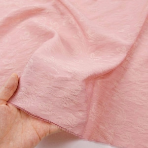 May include: A light pink fabric featuring a subtle, tonal floral jacquard pattern with a textured, slightly crinkled surface. The material is lightweight and semi-sheer, displaying delicate rose motifs woven directly into the textile weave.