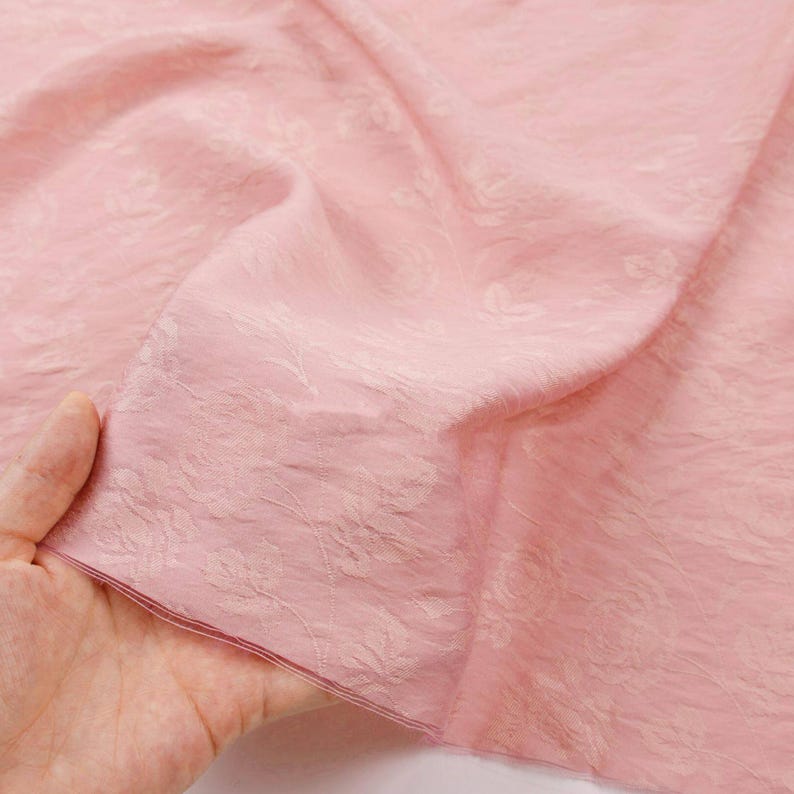 May include: A light pink fabric featuring a subtle, tonal floral jacquard pattern with a textured, slightly crinkled surface. The material is lightweight and semi-sheer, displaying delicate rose motifs woven directly into the textile weave.