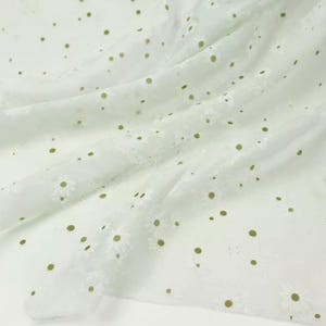 May include: A sheer white fabric featuring an embroidered pattern of small daisies with olive green centers. The lightweight material is draped in soft folds, showcasing the repeating floral design and the translucent quality of the textile.
