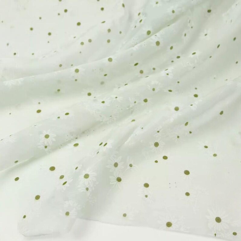 May include: A sheer white fabric featuring an embroidered pattern of small daisies with olive green centers. The lightweight material is draped in soft folds, showcasing the repeating floral design and the translucent quality of the textile.