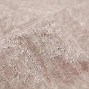 May include: Close-up of a folded piece of fabric in a light beige color. The fabric has a textured pattern of small, repeating geometric shapes. The material appears soft and slightly shiny, with subtle variations in tone.